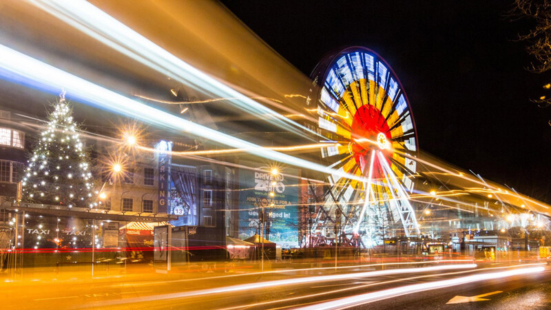 Illuminated Ferris wheel and Christmas tree at night with light trails from passing traffic.