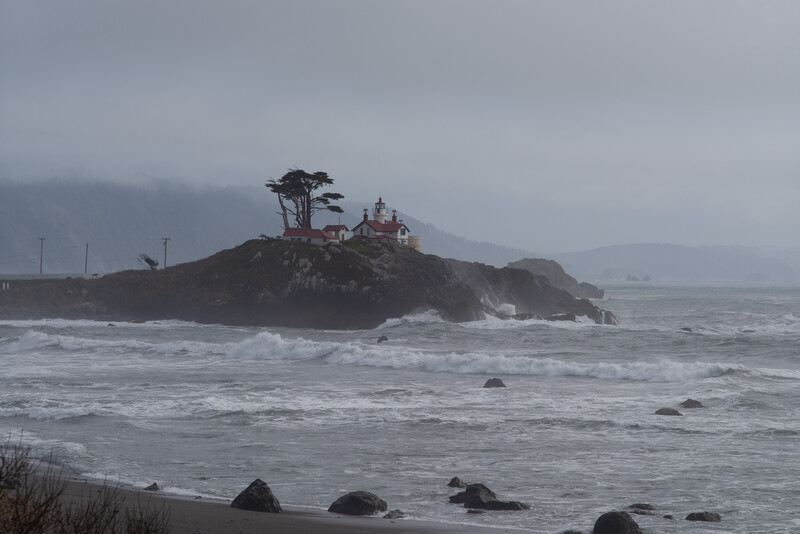 Rocky coastal island with a solitary windswept tree and building, surrounded by rough ocean waves under overcast sky.