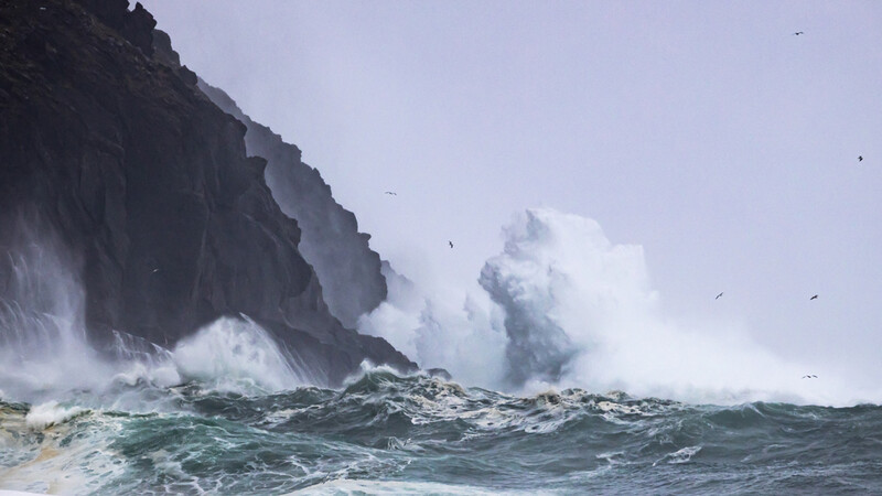 Massive waves crash against dark rocky cliffs under overcast skies.
