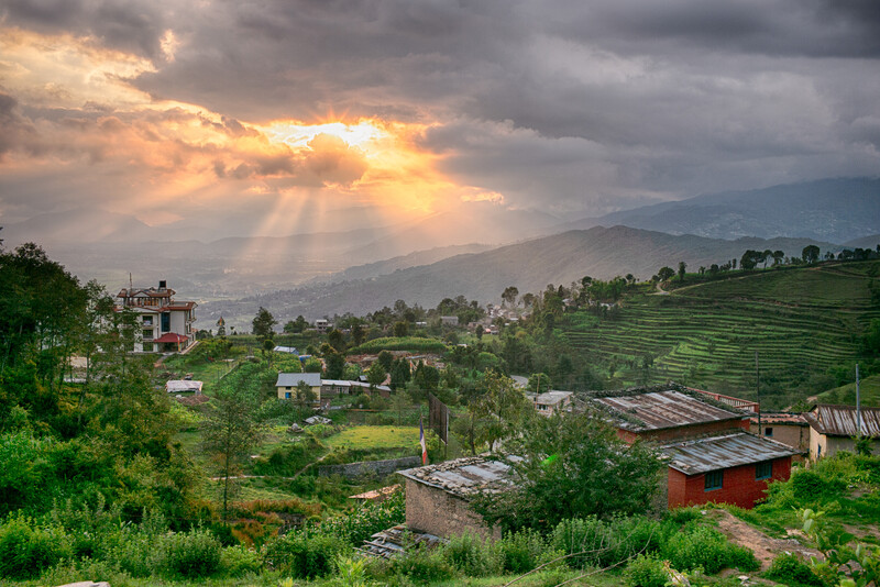 Mountain valley landscape with terraced fields, scattered buildings, and dramatic crepuscular rays breaking through clouds at sunset.