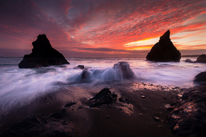 Dramatic sunset over a rocky coastal landscape with silhouetted sea stacks and waves washing across the foreground.
