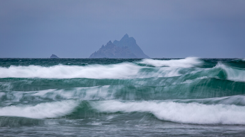 Ocean waves with white foam rolling toward shore, rocky peak visible on misty horizon.