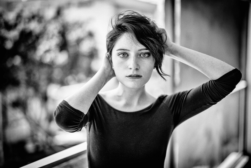 Woman in black shirt with hands near head, gazing directly at camera in black and white portrait.