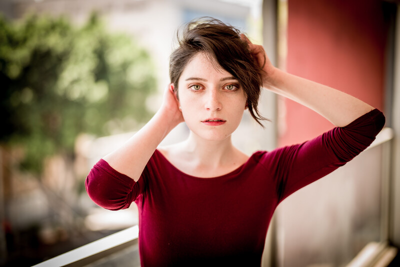 Woman in burgundy shirt with hand in hair, looking at camera with soft natural lighting.