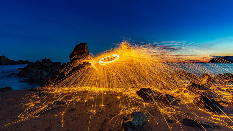 Steel wool spinning creates golden light trails radiating across a rocky beach at twilight.