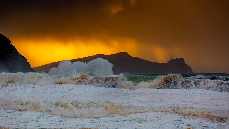 Dramatic golden sunset behind silhouetted coastal cliffs with foaming waves crashing on a sandy beach.