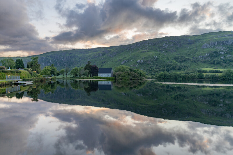 Calm lake reflects dramatic clouds and rolling green hills with scattered buildings and trees.