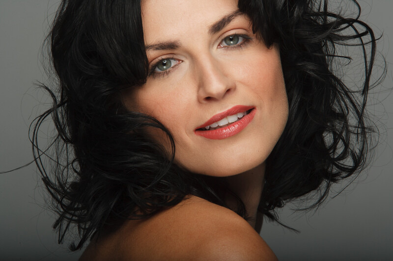 Woman with shoulder-length black wavy hair and red lipstick against a neutral gray background.