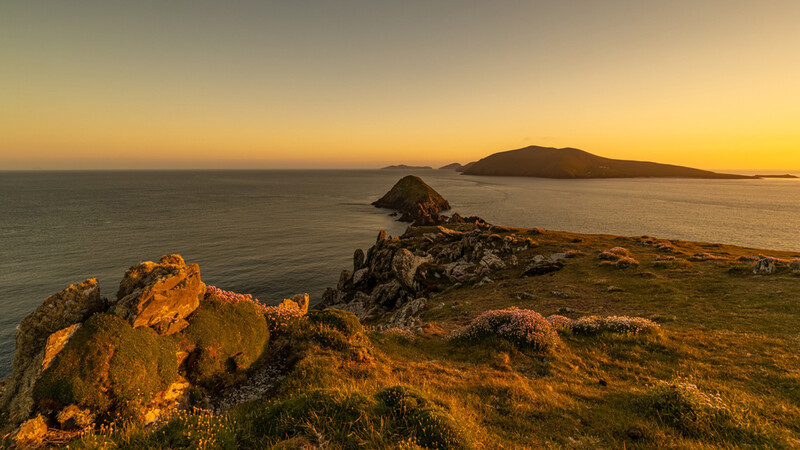 Rocky coastline at golden hour with distant islands silhouetted against sunset sky.