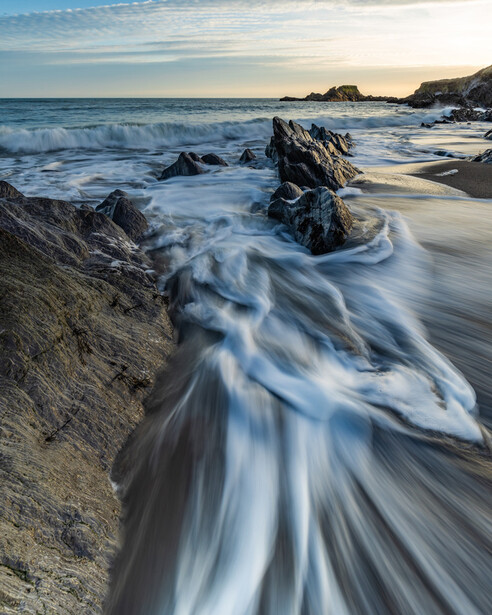 Silky seawater swirls around rocky outcrops on a sandy beach at golden hour.