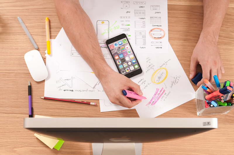 Designer's workspace with wireframe sketches, smartphone, and creative tools arranged on wooden desk.