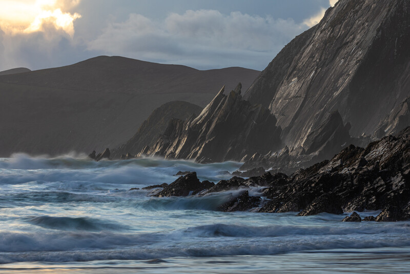 Dramatic coastal scene with towering columnar rock formations, rolling waves, and mountains under cloudy sky.