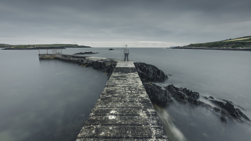 Long stone pier extending into calm gray water with rocky outcrops and distant coastline under overcast sky.