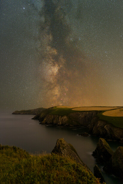 Milky Way galaxy stretching across night sky above coastal cliffs and dark water.