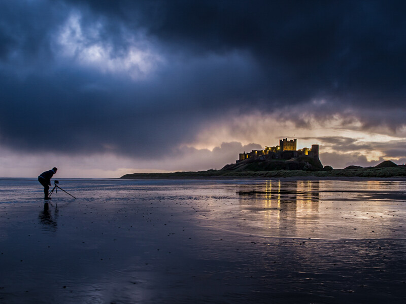 Person wading in shallow water toward a distant castle on an island under dramatic storm clouds.