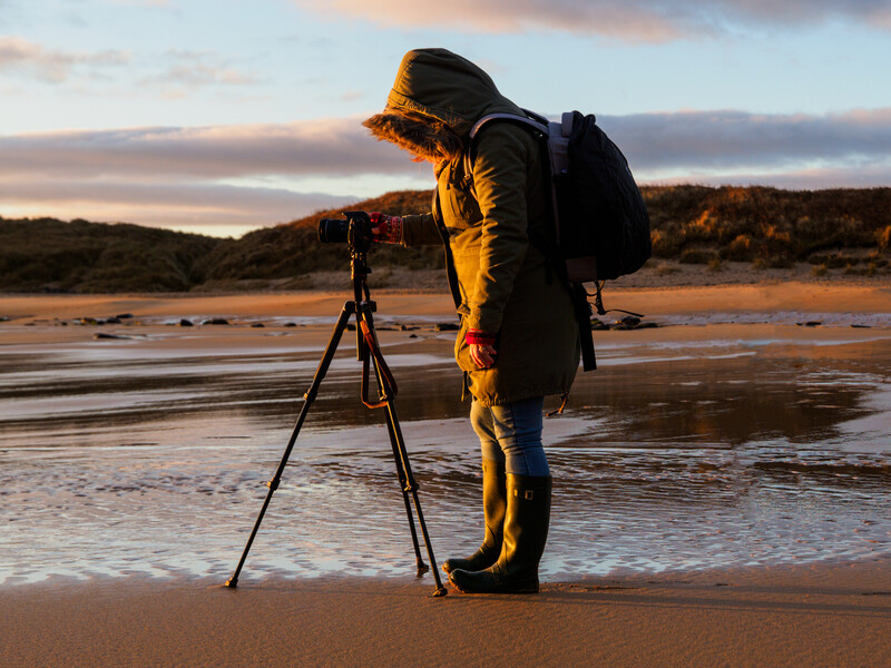 Photographer in yellow jacket using tripod-mounted camera on beach at golden hour.