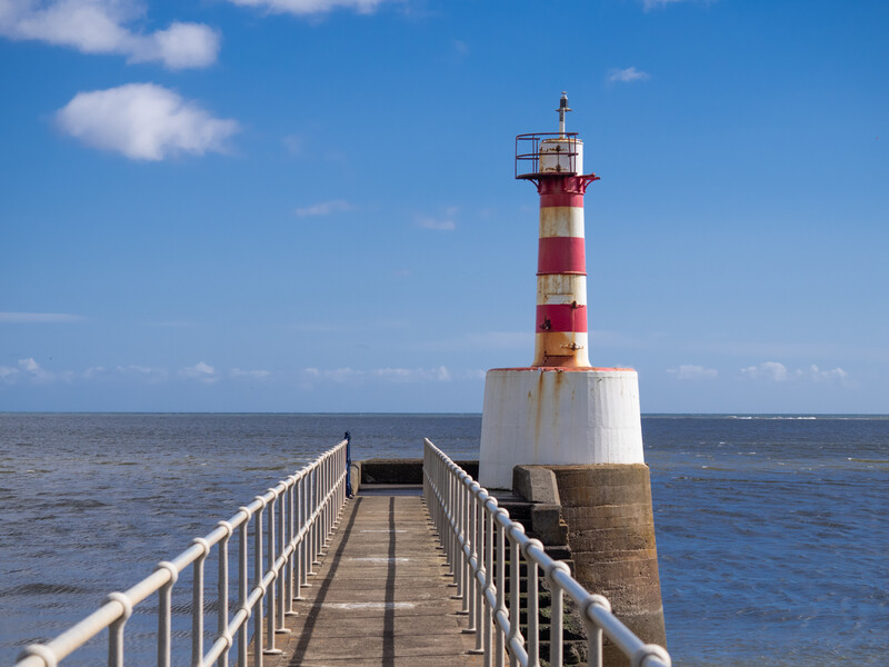 Red and white striped lighthouse at the end of a long white metal pier extending into calm blue sea.