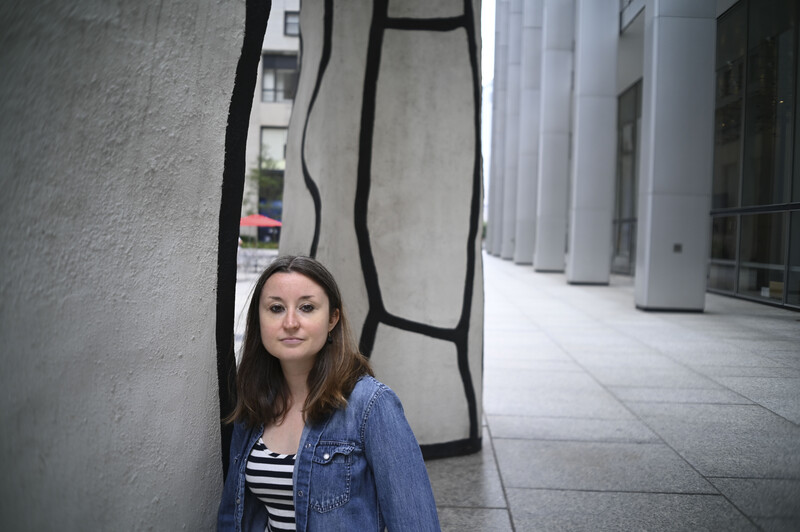 Woman in denim jacket and striped shirt standing against textured wall with black curved lines.