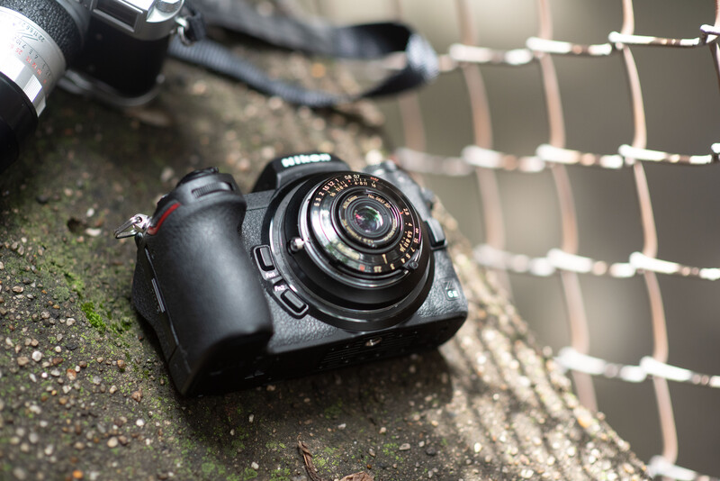 Vintage rangefinder camera with manual focus lens resting on weathered concrete near a wire mesh fence.