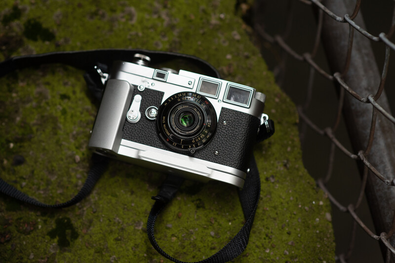 Vintage rangefinder camera with chrome and black body, black strap, positioned on moss-covered ground next to brick.