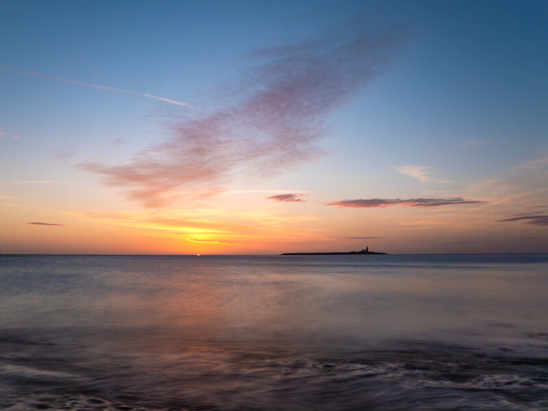 Ocean horizon at sunset with golden and pink clouds reflected in calm water and wet sand.