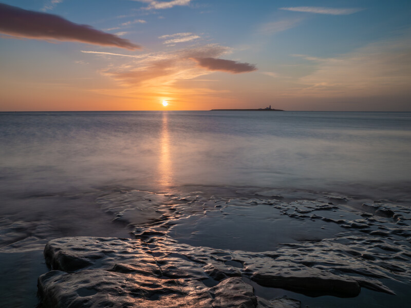 Ocean sunrise with golden light reflecting on calm water and dark rocks in foreground.