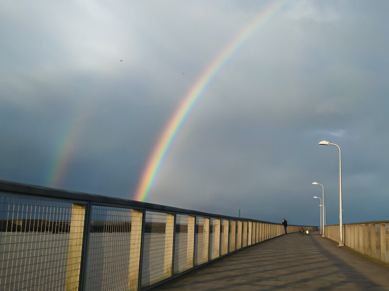 Double rainbow arcing over a bridge with metal railings and street lamps under dramatic cloudy sky.