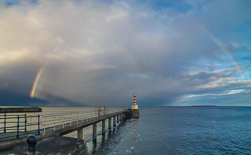 Wooden pier extending into calm blue water with a rainbow arcing from storm clouds overhead.