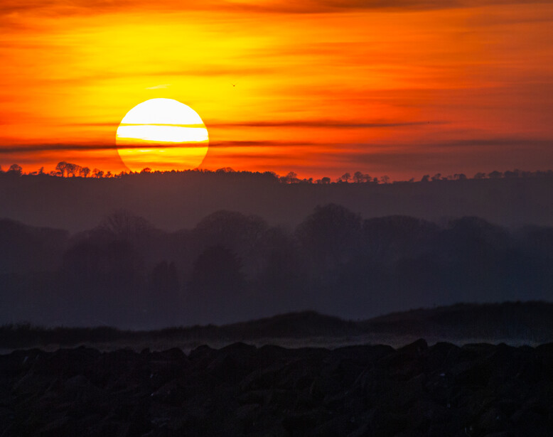 Large sun descending toward a dark, layered landscape at sunset with golden and orange sky.