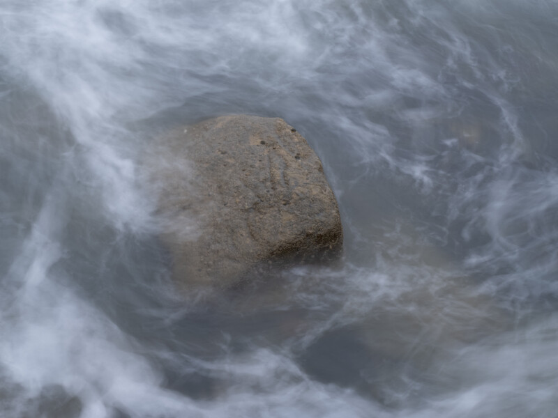 Large rounded boulder surrounded by turbulent white water and foam in shallow ocean waves.