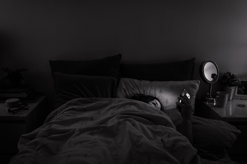 Person lying in bed under dark bedding with a ring light positioned on the nightstand.