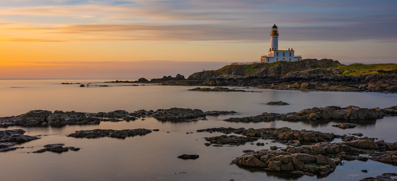 Lighthouse on rocky headland at golden hour with calm waters and scattered rocks in foreground.