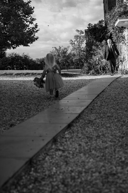 Woman in long dress walking away on a gravel path toward bare trees in an overcast landscape.