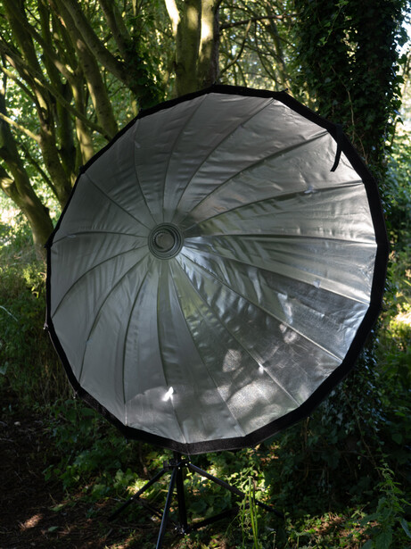 Large parabolic reflector with translucent white fabric diffusion mounted on stand among green foliage.