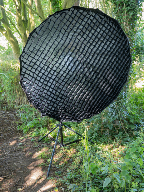 Black mesh reflector with tripod stand positioned among green foliage and trees.