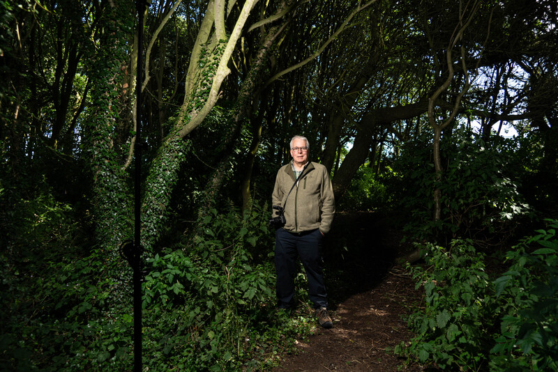 Man in tan jacket standing on a forest path surrounded by moss-covered trees and dense green foliage.