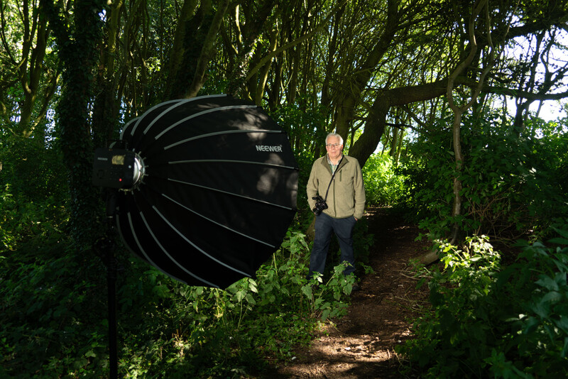 Photographer holding large black reflector dish in shaded forest setting.