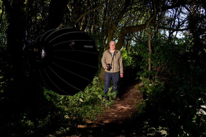 Portrait photographer in khaki vest standing on woodland path holding large reflector disk.