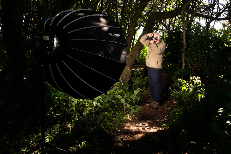 Photographer using a large black reflector to redirect sunlight in a forest setting.