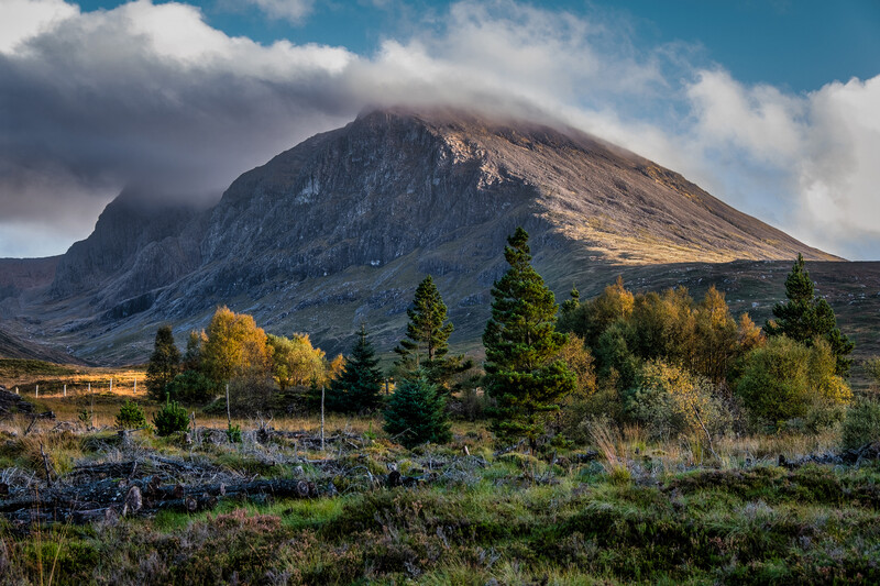 Conical mountain with steep rocky slopes and autumn trees in the foreground under partly cloudy sky.