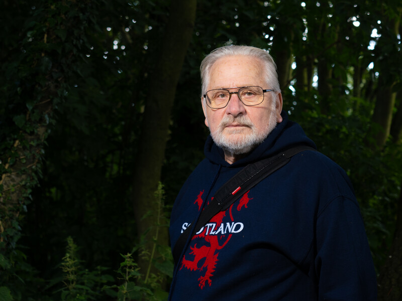 Older man with white hair and glasses wearing a dark navy hoodie with red text, photographed outdoors against evergreen trees.