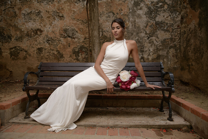 Woman in white sleeveless gown reclining on wooden bench holding red bouquet against weathered stone wall.