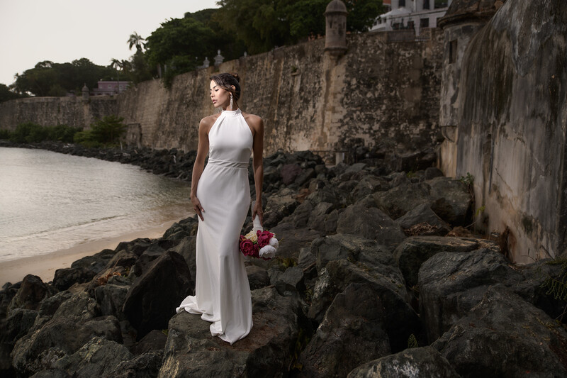 Woman in white wedding dress standing on rocky shoreline against historic stone fortification wall.
