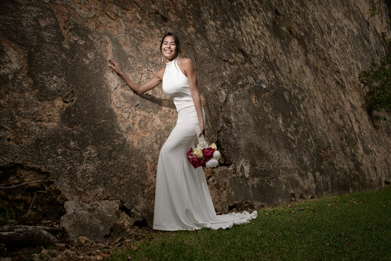 Bride in white halter gown holding red and pink bouquet against weathered stone wall.
