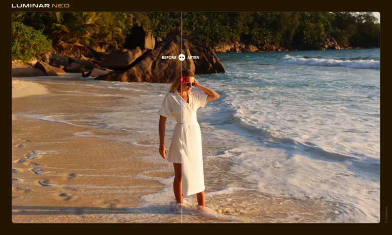 Woman in white dress standing in shallow ocean water on a sandy beach with waves and forested shoreline behind her.