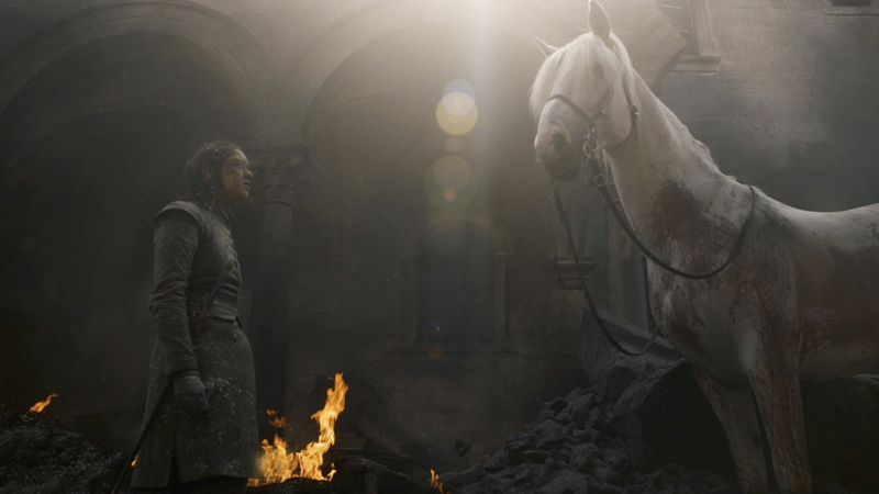Person in medieval clothing facing a white horse in a stone archway lit by firelight and golden light.