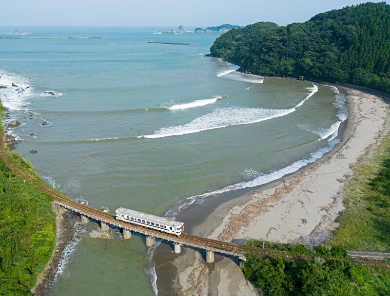 Aerial view of a curved beach with a train crossing a bridge over a river mouth, forested hillside beyond.