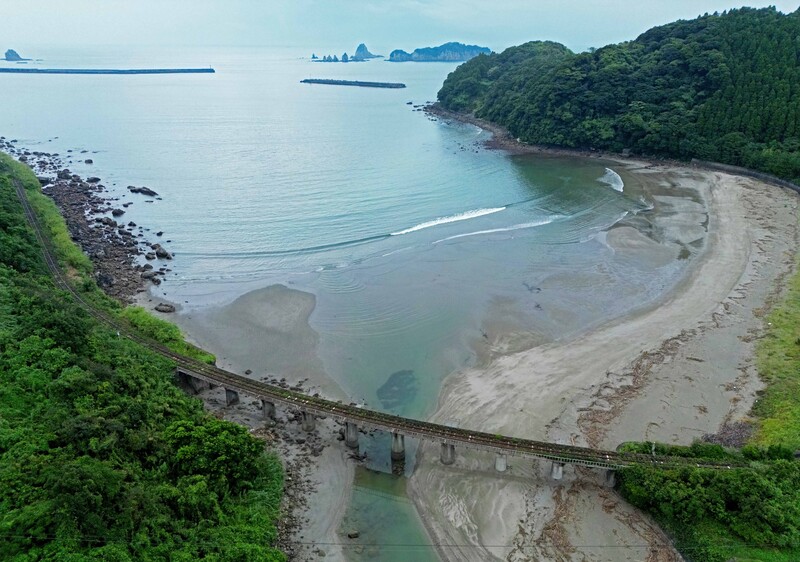 Aerial view of a curved sandy beach cove bordered by dense green forest and calm turquoise water.