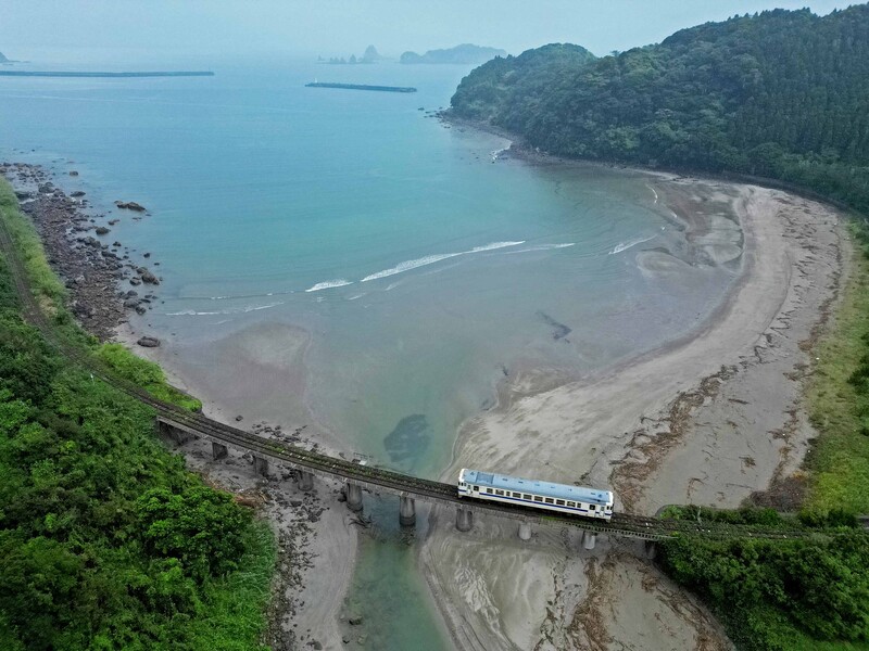 Elevated view of a passenger train crossing a bridge over a curved beach with forested headlands.