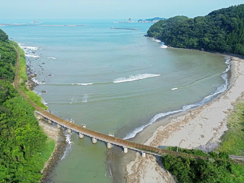 Aerial view of a curved coastal beach with a bridge crossing a river mouth, dense green forest, and turquoise ocean.